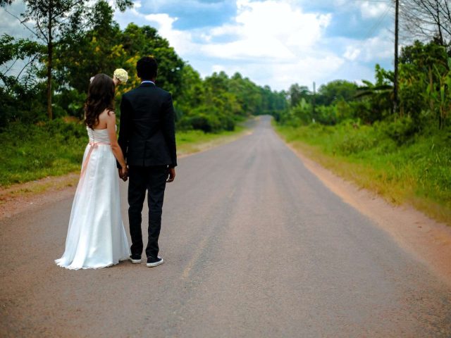 wedding couple standing on winding road between trees under cumulus cloud